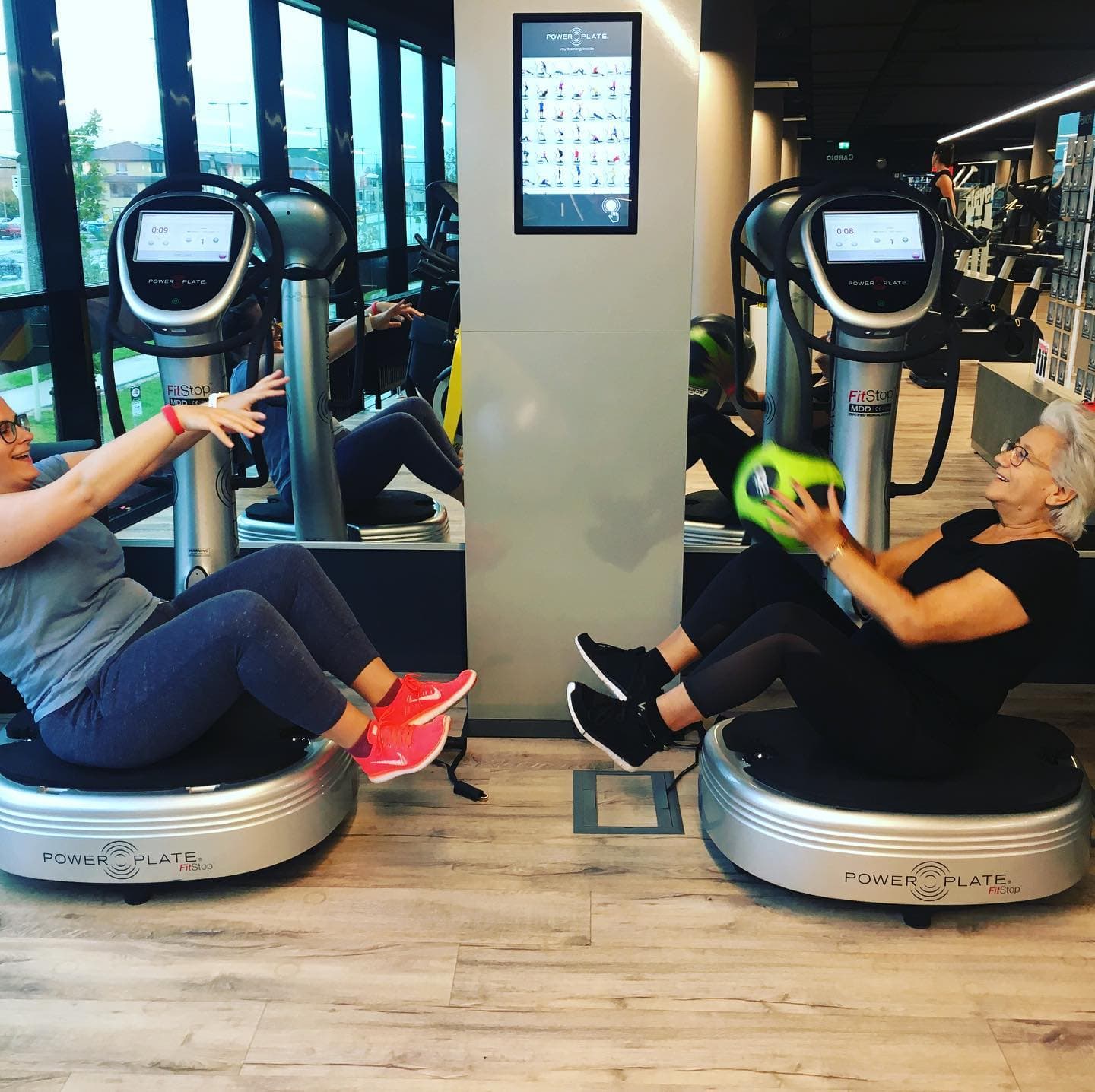 Vibration Plate Workout in Gym Two women doing seated exercises on vibration plate machines in a gym.